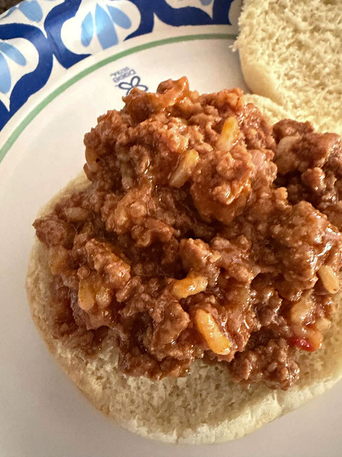 An open-face sloppy joe sandwich with the top bun to the right on a paper plate.