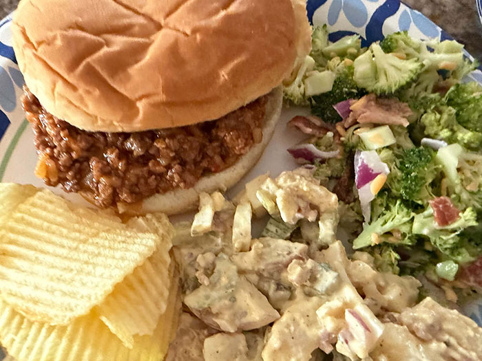 A sloppy joe sandwich on a paper plate with a serving of broccoli salad, potato salad, and ruffled potato chips. 