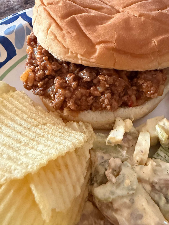 A sloppy joe sandwich on a paper plate with ruffled potato chips and potato salad.