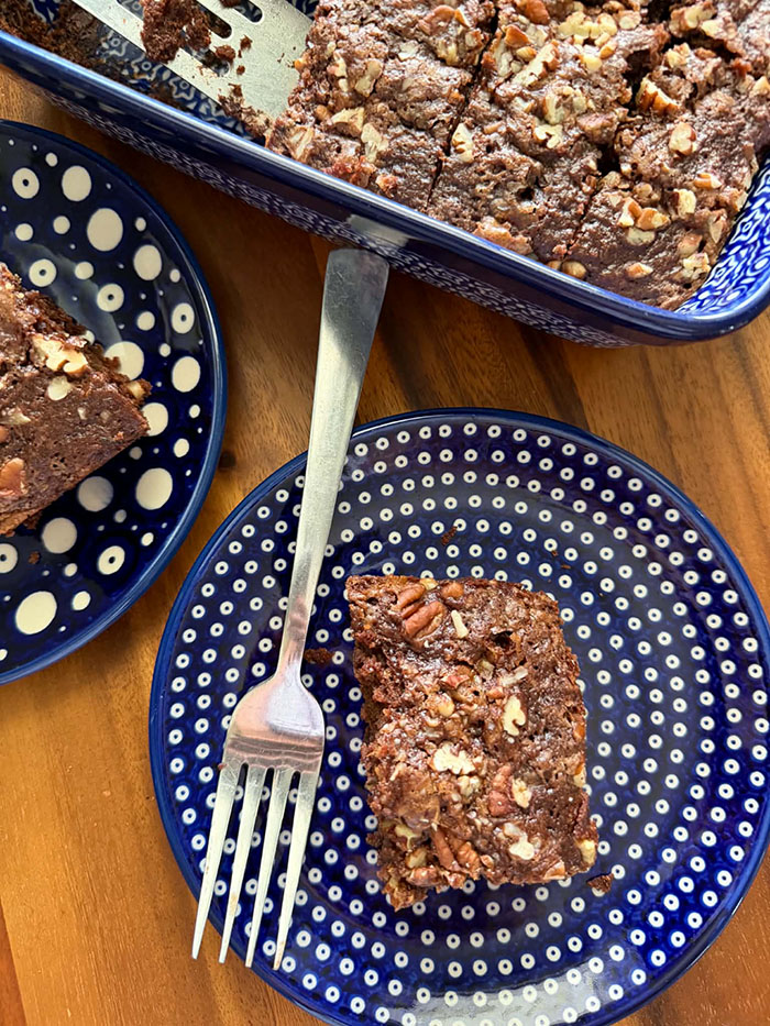A corner piece of zucchini chocolate cake with pecan streusel on a small dark blue plate with polka dots. Fork is lying on the plate on the left of the piece of cake. A partial picture of a small dark blue plate with large and small polka dots with a slice of zucchini chocolate cake with pecan streusel is in the upper left. The corner of a dark blue 9x13 baking dish of sliced chocolate zucchini cake with pecan streusel is at the top middle and right.