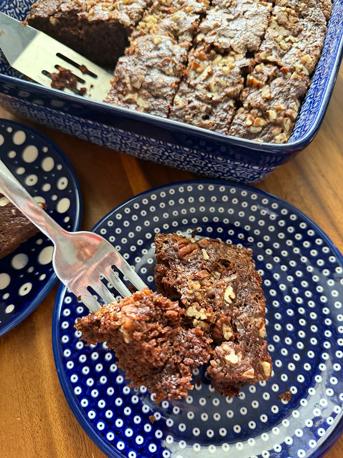 A slice of zucchini chocolate cake with pecan streusel on a small dark blue plate with white polka dots with a fork held above with a bite of cake on it. At the top is a 9x13 baking dish of sliced chocolate zucchini cake with pecan streusel with a serving spatula inserted where a missing piece of cake was.
